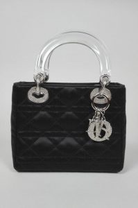 CHRISTIAN DIOR, PARIS. Modèle : Lady Dior Mini. Sac à main à deux poignets en satin noir surpiqué au motif Cannage, les poignets en plastique transparent et les garnitures en métal argenté orné de strass cristal, fermant par un rabat en satin noir. L'intérieur en toile crème et argent au motif assorti à la surpiqure extérieure, avec un grand compartiment et une petite poche zippée. 17 x 15 x 7 cm. Présenté avec son dustbag et sans bandoulière.